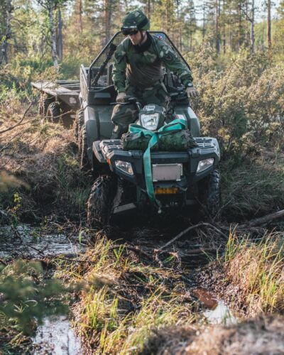Soldat i Fältuniform 90 kör en Terränghjuling 6x6 med släpvagn genom ett vattendrag i skogsterräng.